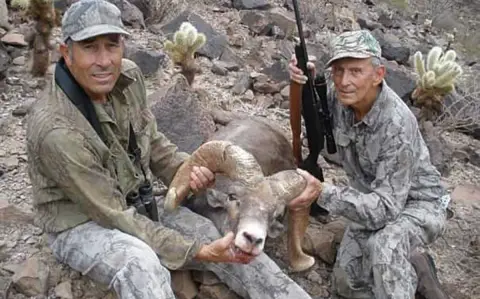 Deux hommes en tenue de camouflage tiennent un mouflon d'Amérique par ses cornes tout en étant assis sur le sol rocheux