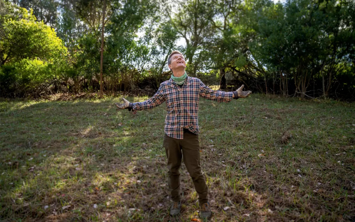 Ole Schell se tient dans un champ herbeux avec une chemise à carreaux et les bras écartés, souriant et regardant le ciel.