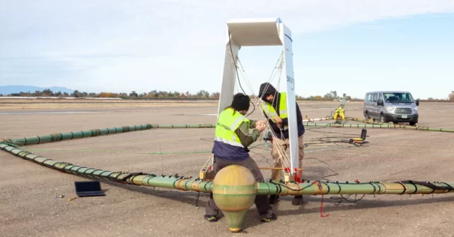 Des travailleurs en tenue réfléchissante effectuent des ajustements sur une grande pièce d'équipement de forme géométrique sur un aérodrome.