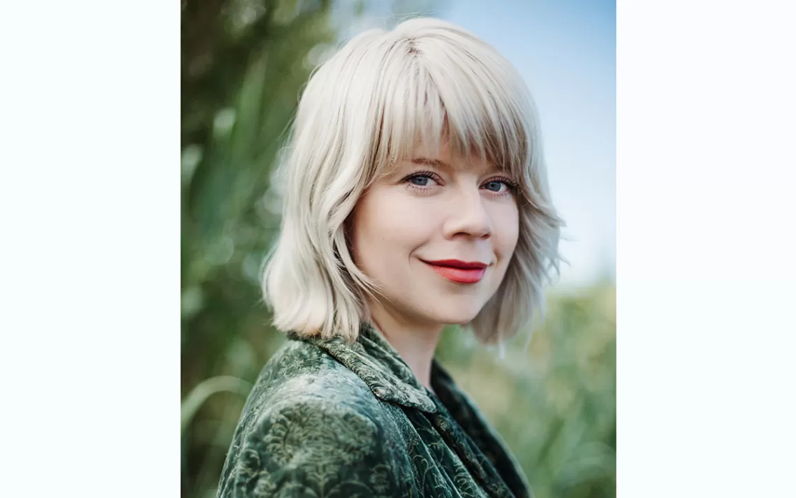 Personne en robe verte et cheveux blonds debout devant la caméra