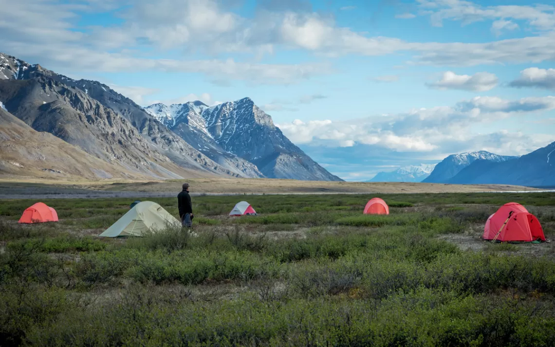   Un homme se tient près de quatre tentes dans une prairie de la vallée de l'Arctic Refuge avec des montagnes aux sommets enneigés en arrière-plan.