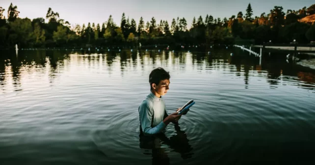 Un jeune homme se tient debout au lac Temescal, à Oakland, au crépuscule, tenant un iPad.  Il y a une douce lueur sur son visage.