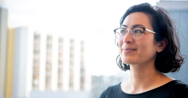 Rose Abramoff porte des lunettes et un haut noir et sourit en regardant à sa droite.