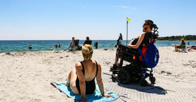 Une personne est assise dans un fauteuil roulant sur la plage, à côté d’une autre personne assise sur une serviette sur le sable.
